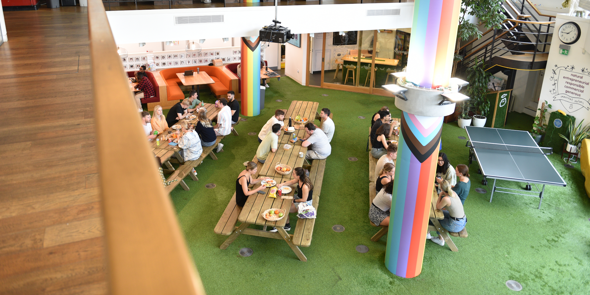 People eating at picnic tables with a rainbow pillar in an office with a ping pong table.