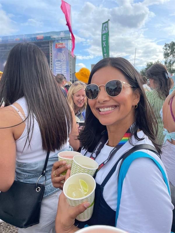 Woman with sunglasses, holding drinks, smiles at camera at a festival.