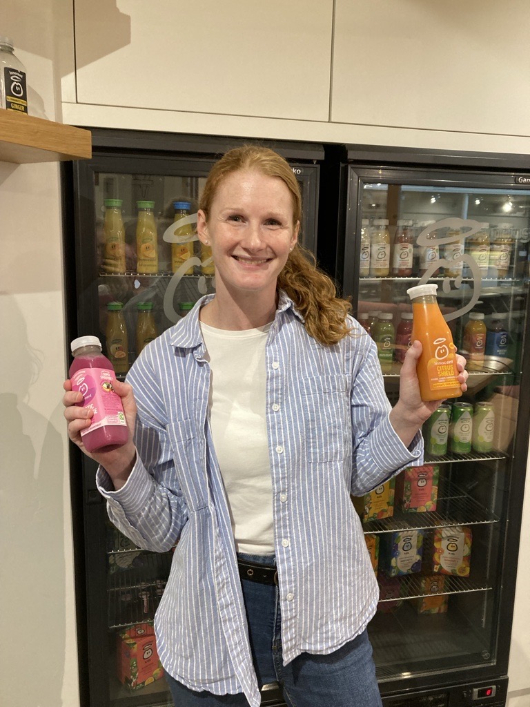 Woman holding two juice bottles, standing in front of a cooler filled with drinks.