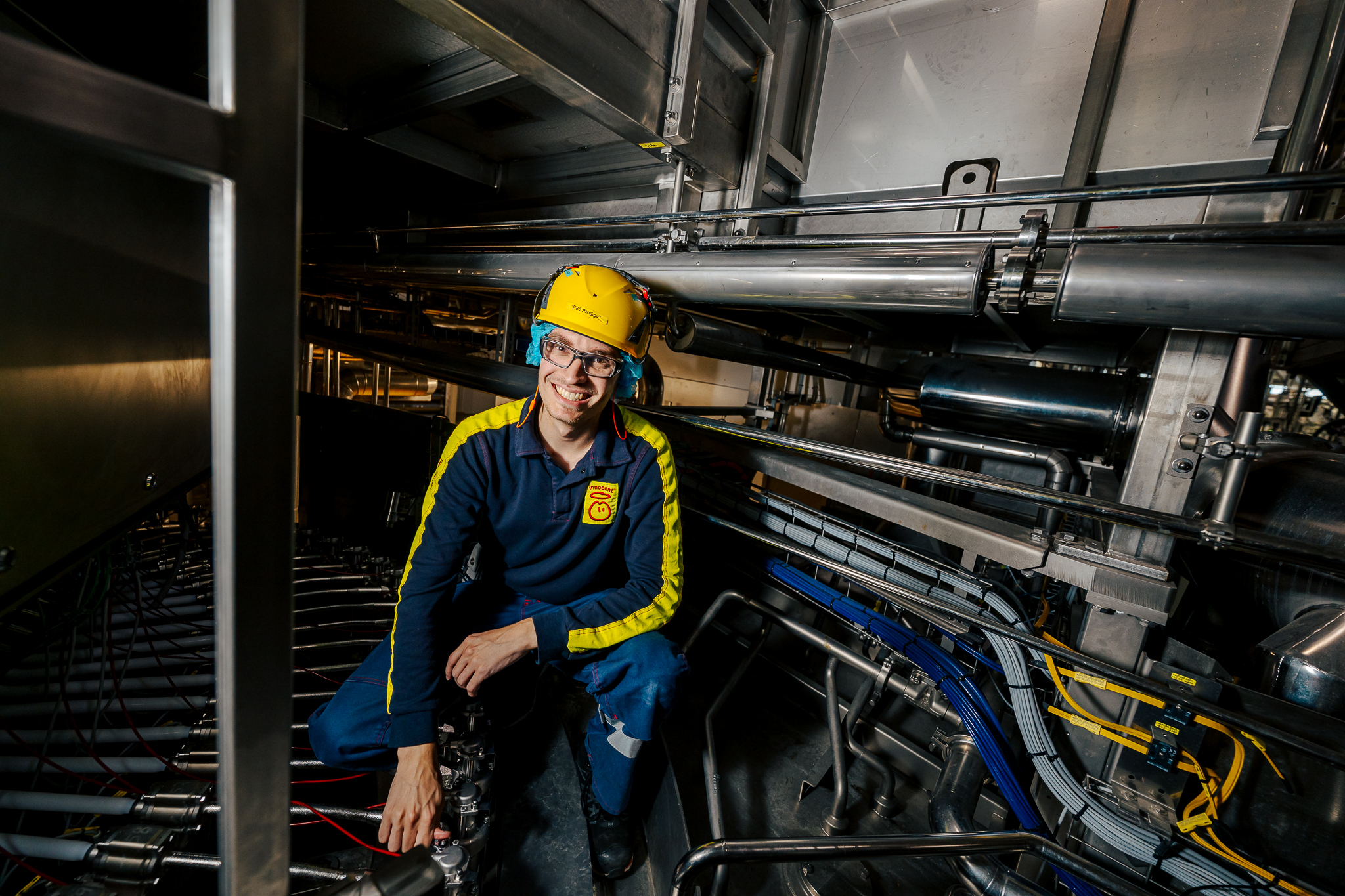 Smiling worker wearing a hard hat, glasses, and uniform, surrounded by machinery.