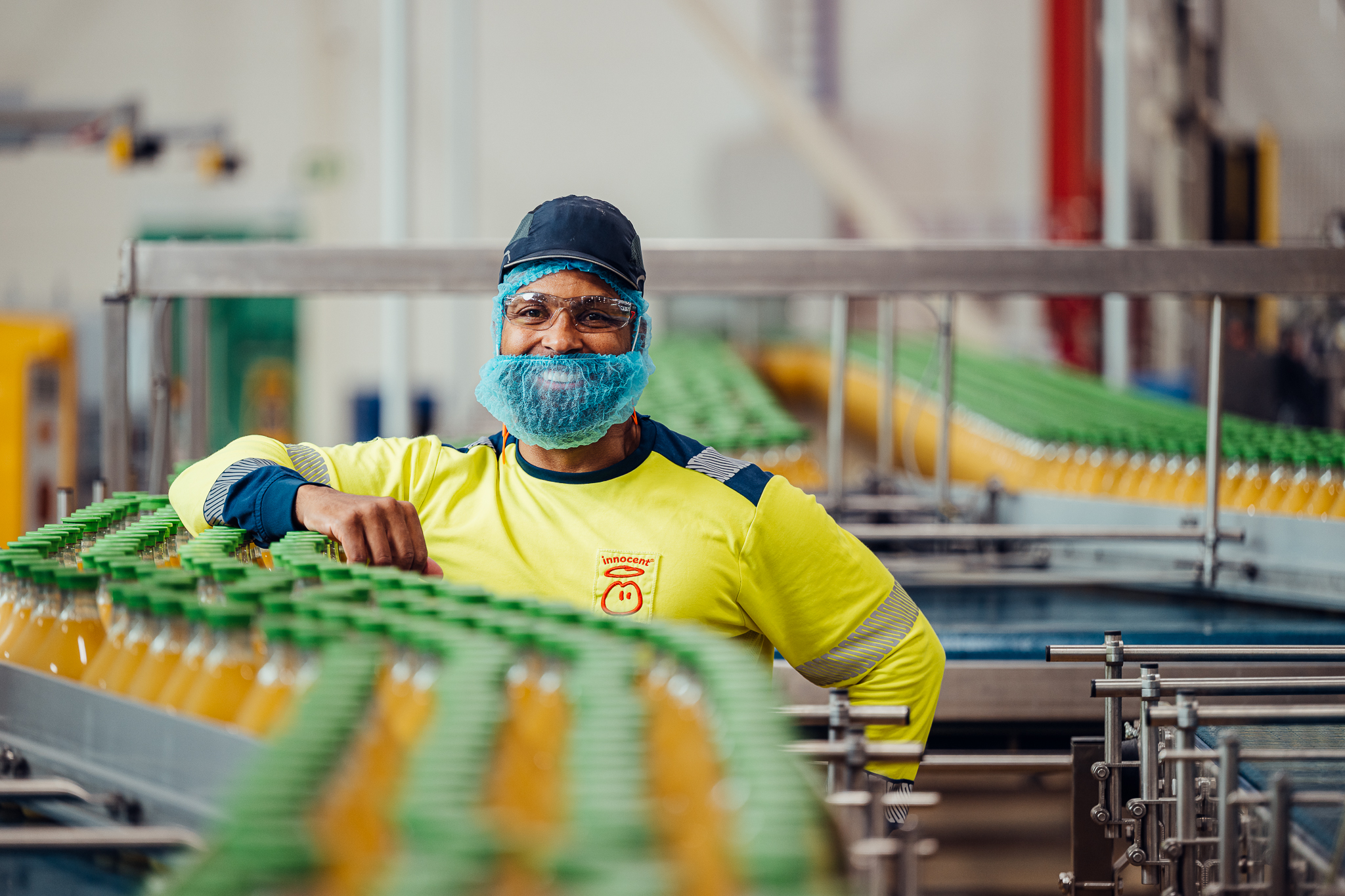 Worker in protective gear by juice bottles on a conveyor belt.