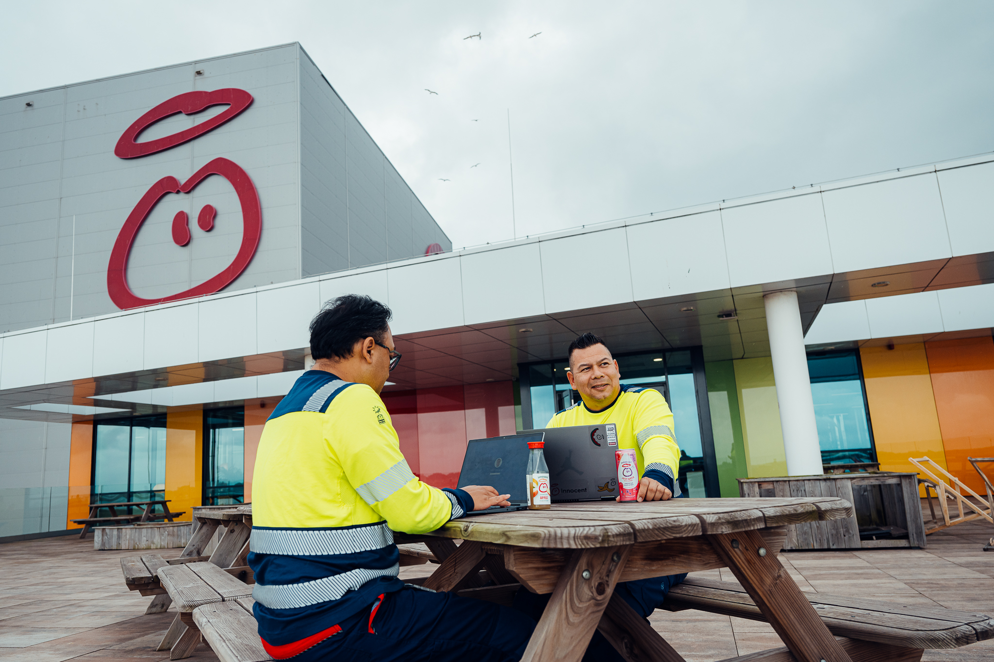 Two men at a table with laptops, Innocent building in background.