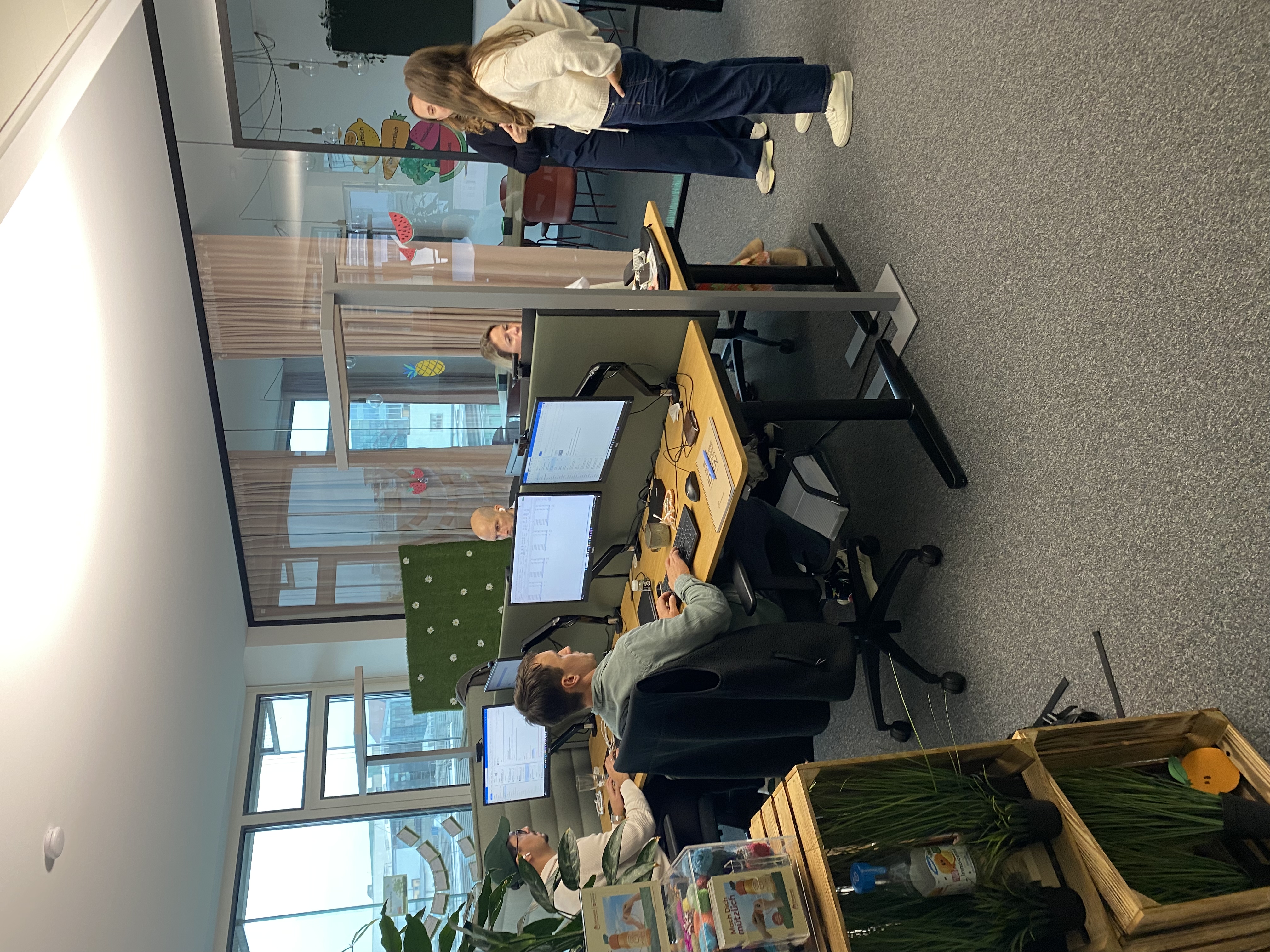 Several people work at desks with computers in a modern office. Glass partitions divide the workspace.